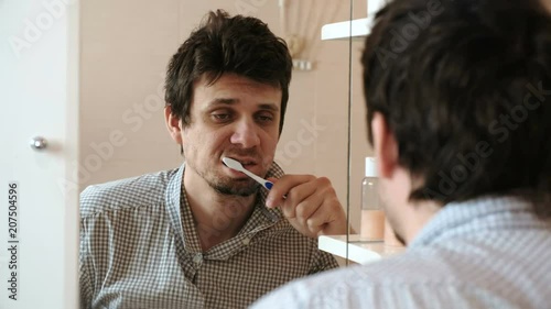 Tired man who has just woken up brush his teeth, looks at his reflection in the mirror and sees his scruffy appearance.