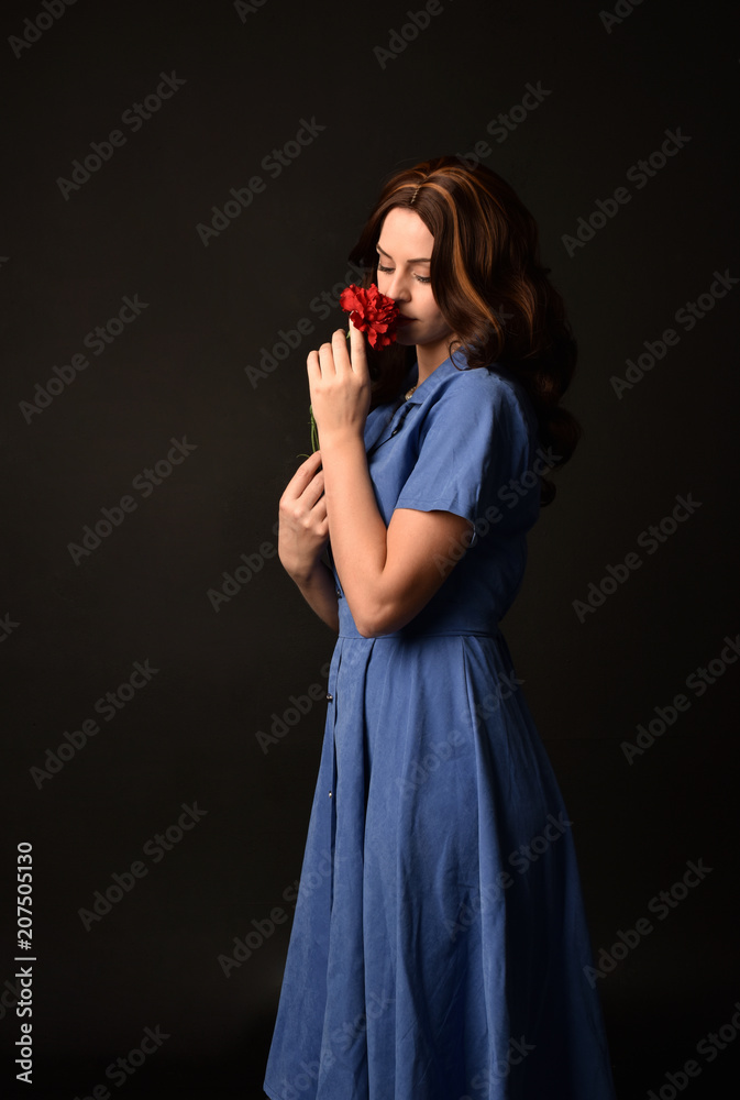 3/4 portrait of brunette lady wearing blue dress holding a red flower. posed on black studio background. 3/4 portrait of brunette lady wearing blue dress holding a red flower. posed on black studio background.