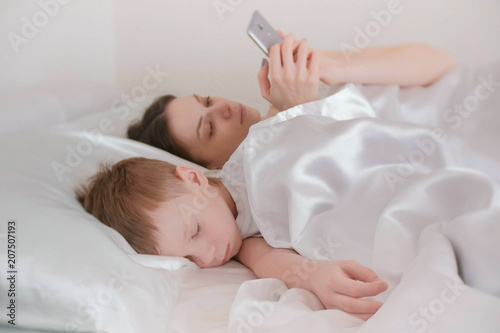 Mom woke up and browsing internet in her mobile phone. Her son is still asleep.