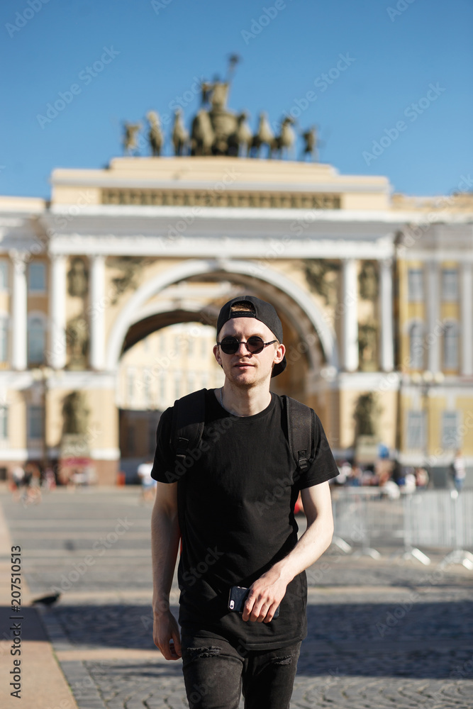 Fototapeta premium Young stylish man tourist walking near General Staff Builsing arch in the palace square in St. Petersburg. He wearing in black jeans, black T-shirt, black cap with riund sunglasses