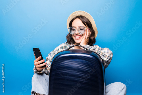 Wallpaper Mural Young pretty woman in sunglasses and straw hat with a suitcase looks into the phone isolated on blue background. Flight traveling. Torontodigital.ca