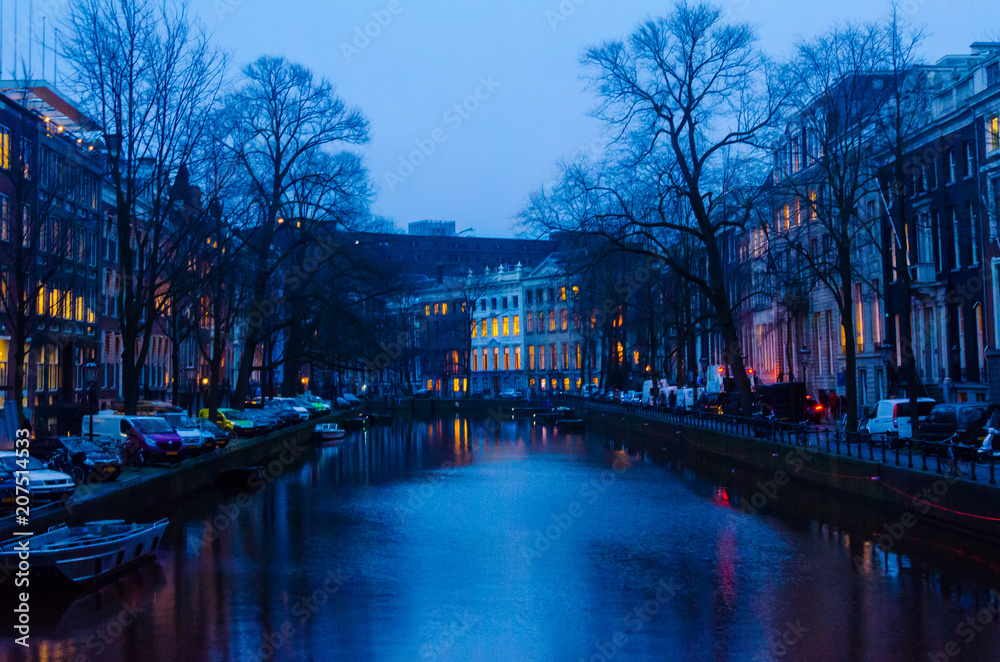Historical old residential house building along river water canal of amsterdam during sunrise with lights and empty street pavement under cloudy warm sky with church background