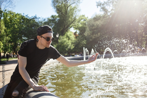 Stylish man at the fountain in hot summer day. Man cooling himself by splashing water He wearing in black jeans, black T-shirt, black cap with riund sunglasses photo