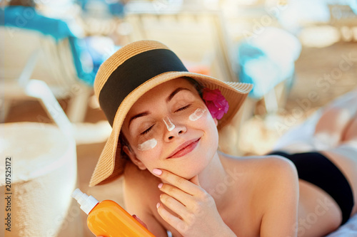 Young woman with sun cream on face holding sunscren bottle on the beach. Female in hat applying  moisturizing lotion on skin.
