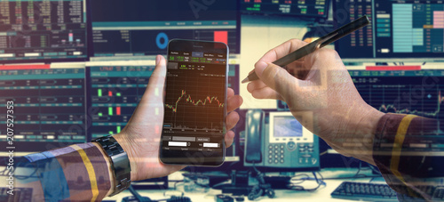 Double exposure of hand holding smartphone with Stock market quotes and chart in monitor computer room with business office equipments .business and money concept