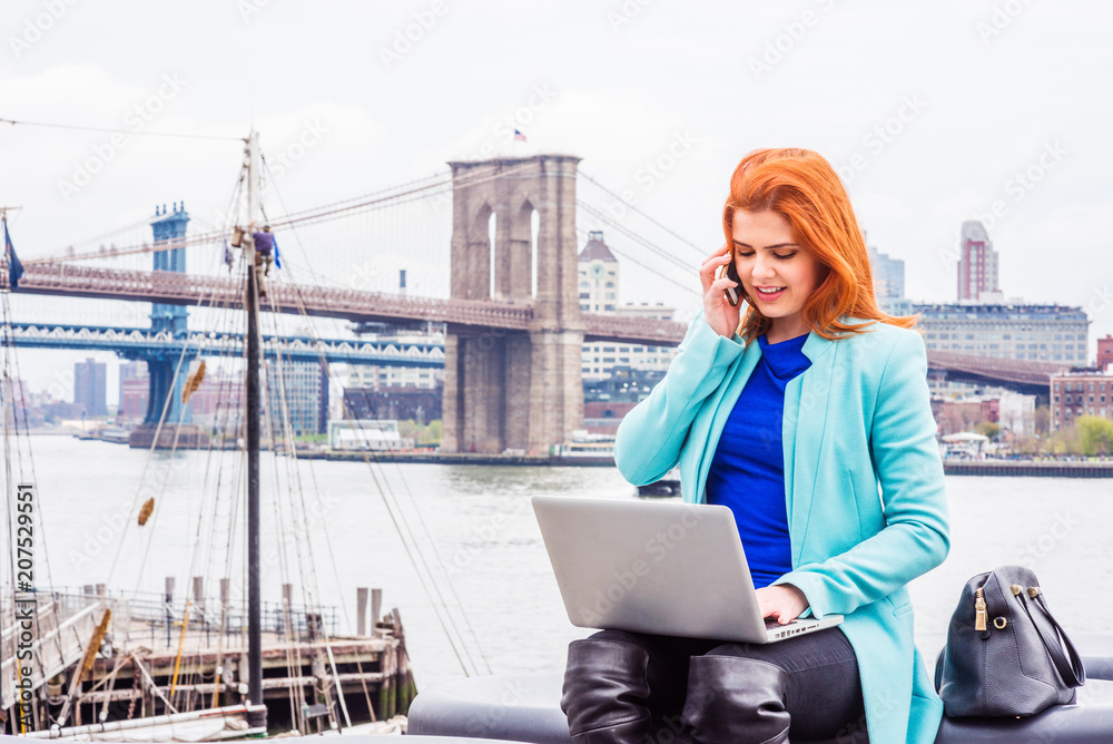 Young American Woman traveling in New York, wearing long cadet blue woolen overcoat, sitting by river, working on laptop computer, calling on cell phone. Manhattan, Brooklyn bridges on background..
