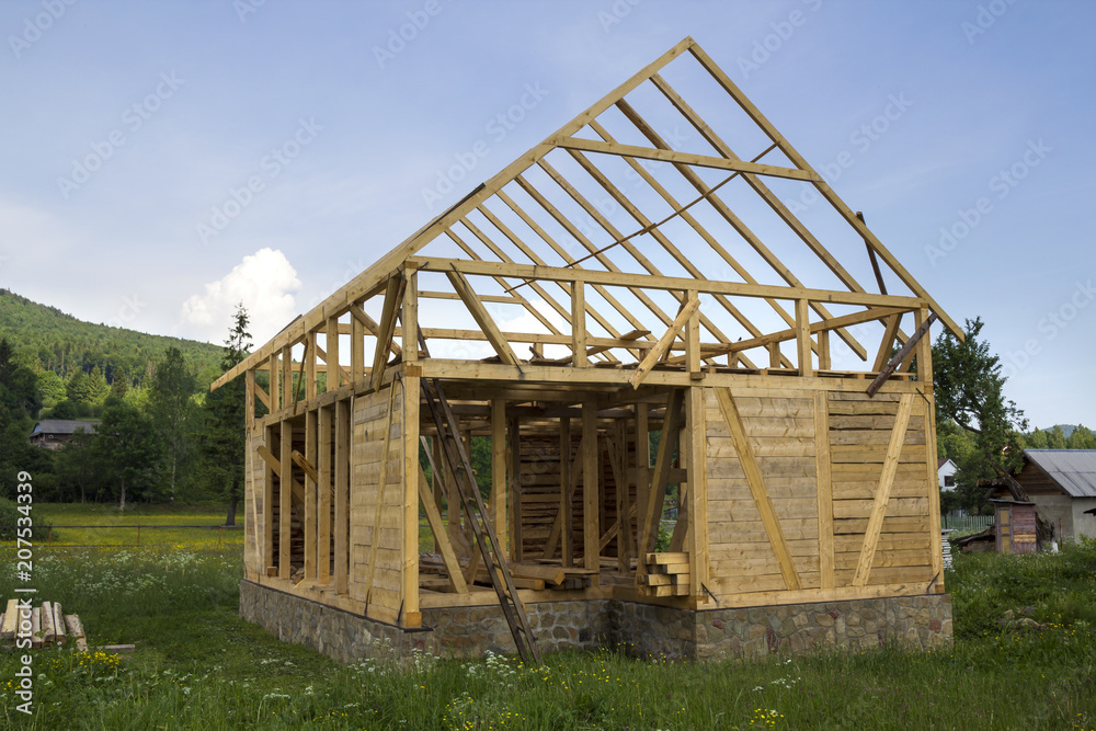 New wooden house under construction in quiet rural neighborhood. Timber frame of natural materials for walls and roof on stone foundation. Property, professional building and reconstruction concept.