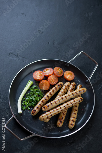Metal pan with grilled pork sausages, tomatoes and green onion, elevated view on a black stone background, copyspace
