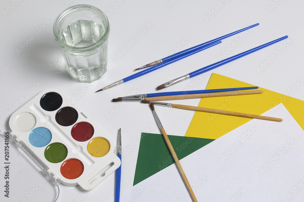 Watercolors and brushes on a white surface. Next glass for water and paper. Subjects for creativity. Watercolors and brushes on a white surface. Next glass for water and paper. Subjects for creativity.
