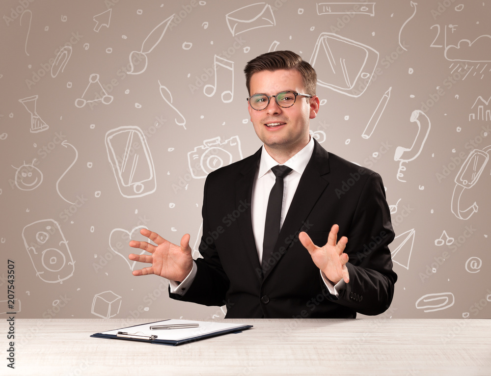 Young handsome businessman sitting at a desk with white mixed media icons behind him Young handsome businessman sitting at a desk with white mixed media icons behind him