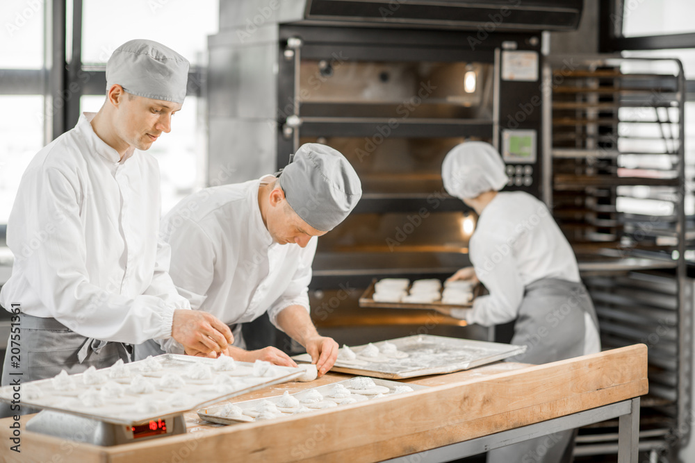 Group of young workers in uniform filling buns for baking on the wooden table standing together at the modern manufacturing Group of young workers in uniform filling buns for baking on the wooden table standing together at the modern manufacturing