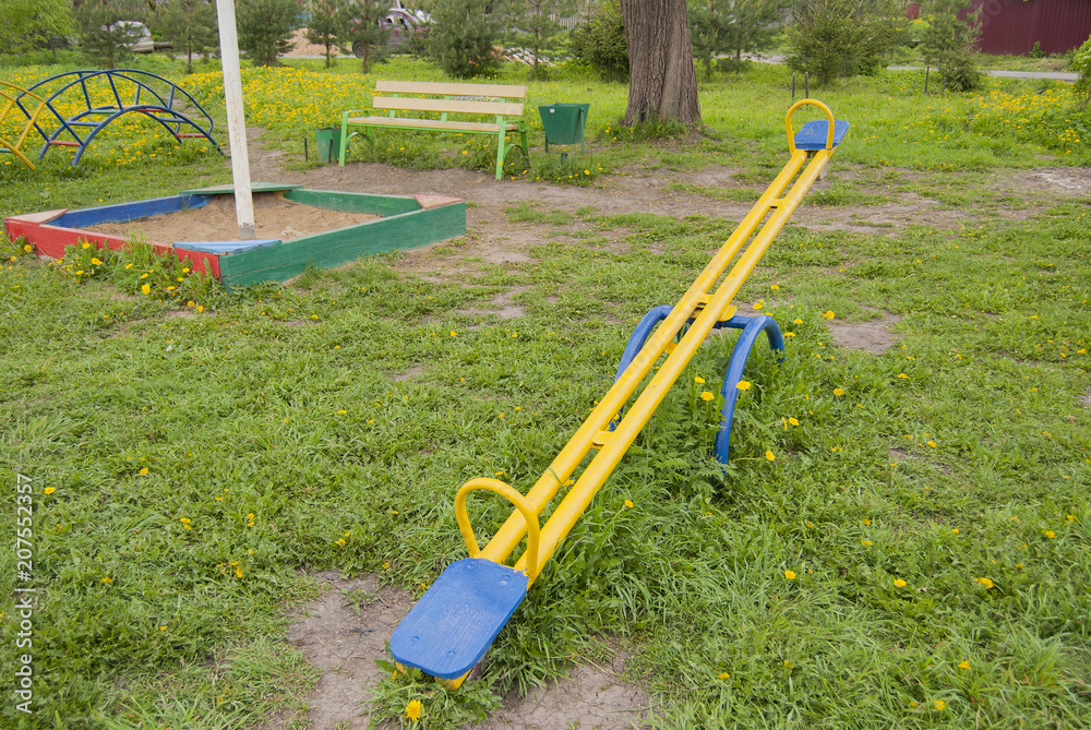 Fototapeta premium Swing for children, yellow with blue seats on a background of green grass with yellow dandelions