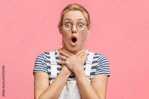 Portrait of stupefied attractive female has attack of coughing, keeps mouth opened and eyes popped out, hands on neck, wears casual t shirt, poses against pink studio wall. People and body language