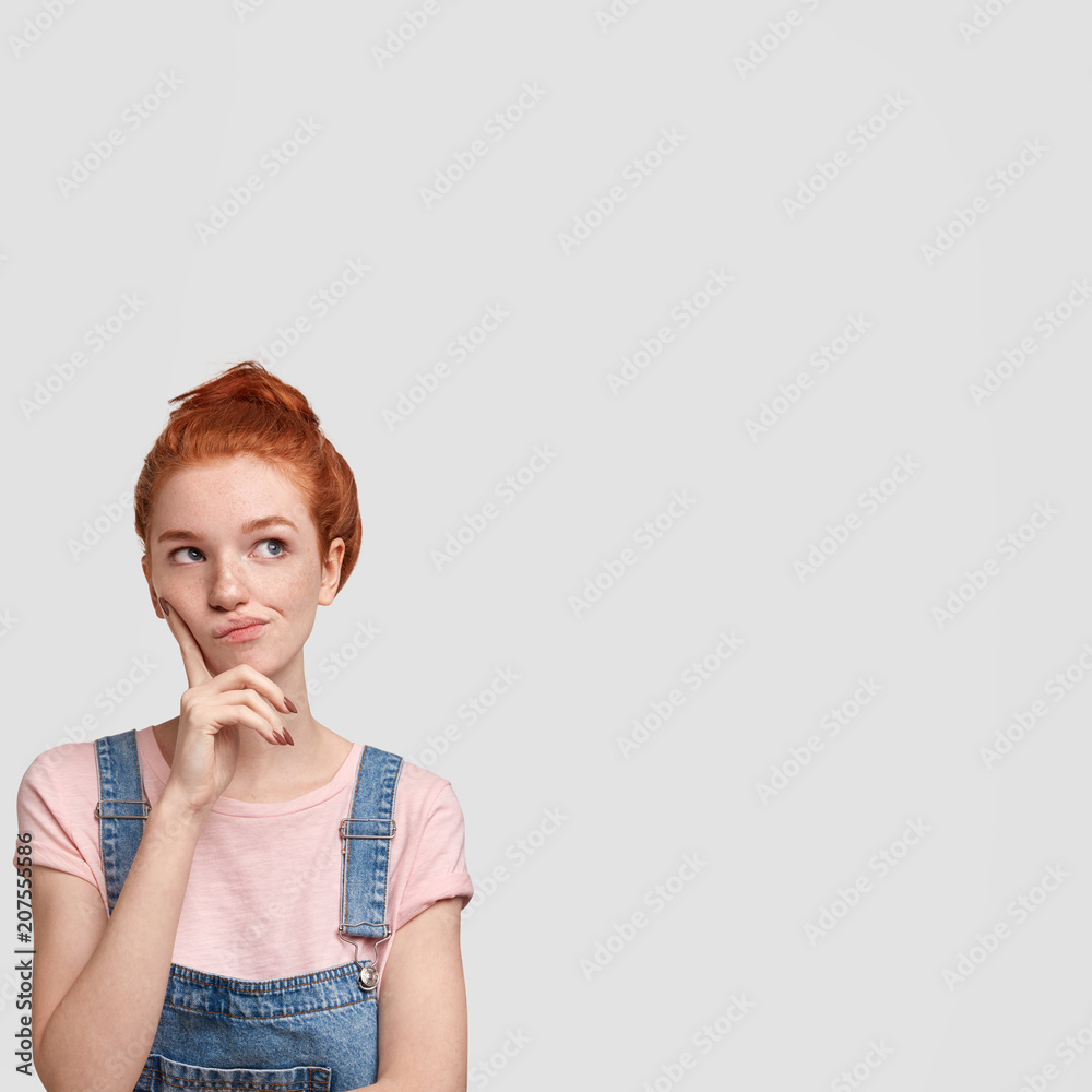 Φωτογραφία Vertical shot of lovely redhead female being deep in thoughts, curves lips and looks pensively upwards, poses against white blank wall with copy space for your advertising content
