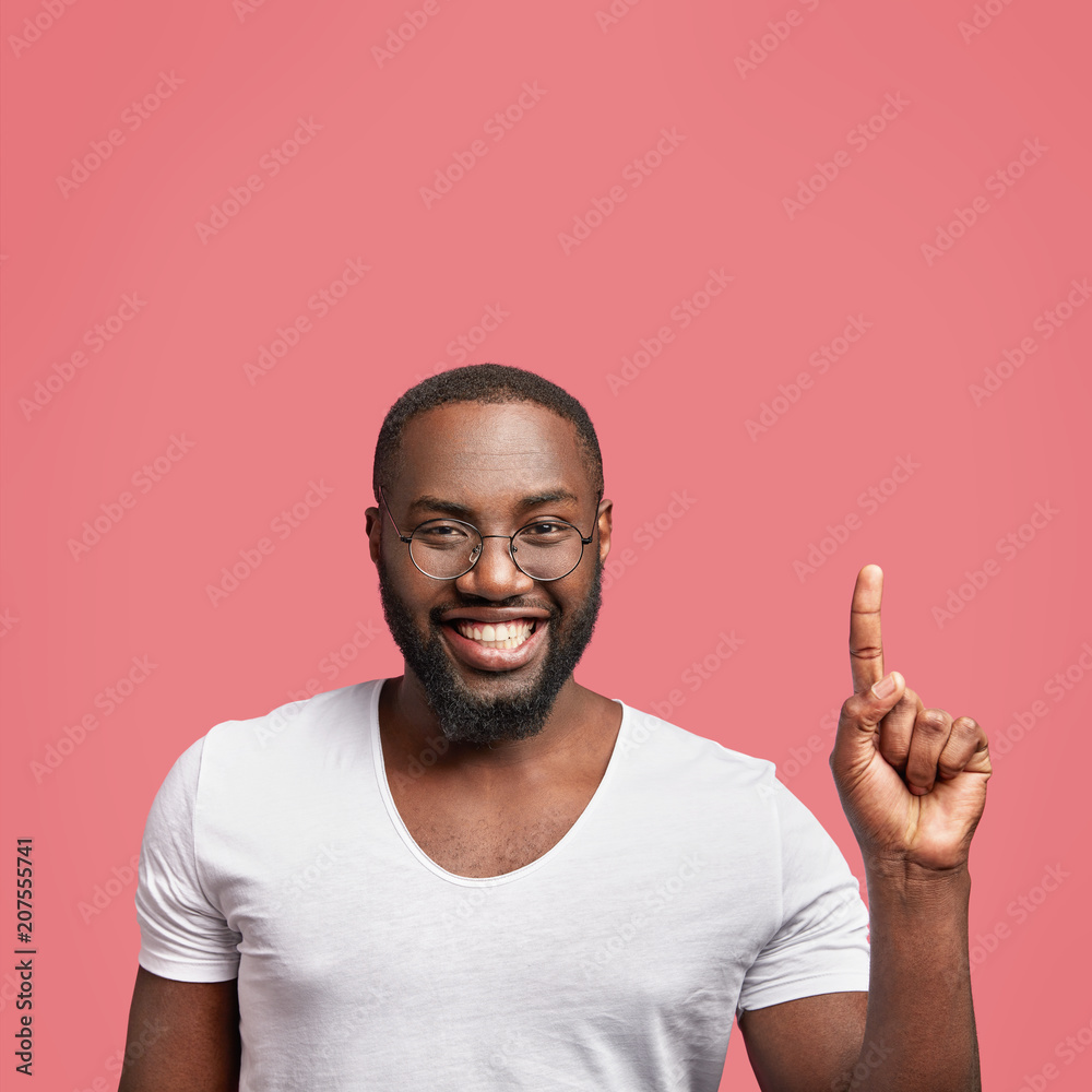 Good looking dark skinned unshaven male with broad smile, has white perfect teeth, raises fore finger upwards, advertises new products of his shop, wears spectacles, isolated over pink background Good looking dark skinned unshaven male with broad smile, has white perfect teeth, raises fore finger upwards, advertises new products of his shop, wears spectacles, isolated over pink background