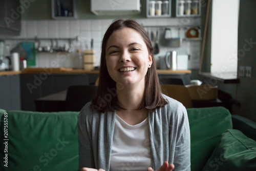 Smiling millennial girl laughing sitting on kitchen sofa talking by videocall looking at camera, female teen video blogger recording vlog at home, casual videoblogging concept, head shot portrait