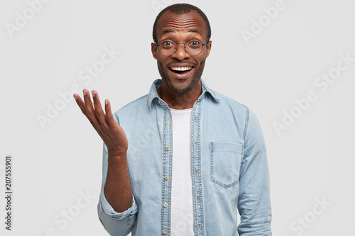 Positive dark skinned African American male giggles as hears something funny, gestures with hand, dressed in casual outfit, has pleasant talk with wife, isolated over white background. Happy young man