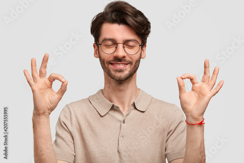 Portrait of happy unshaven young male hipster makes ok sign, has stubble, keeps eyes closed, meditates indoor, poses against white background. Glad Caucasian man gestures indoor and shows approval
