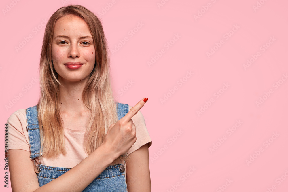Portrait of gorgeous young female model with positive expression, wears casual clothes, indicates with fore finger at blank copy space, shows space for your advertisement or promotional text. Portrait of gorgeous young female model with positive expression, wears casual clothes, indicates with fore finger at blank copy space, shows space for your advertisement or promotional text.