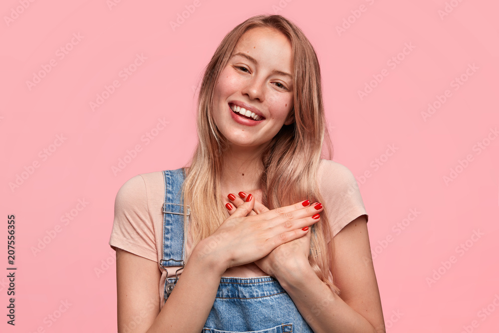 Lovely cheerful female with broad smile and friendly look, keep hands on chest, expresses positive and good attitude, has red manicure, feels gratitude to her mother, isolated on pink background