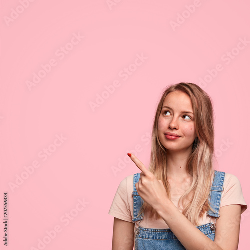 Tableau sur toile Portrait of beautiful woman with European appearance indicates with fore finger at upper left corner, shows blank space for your advertisement, isolated over pink background