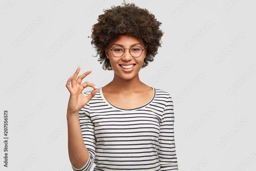 Positive mixed race female model has Afro hairstyle, shows okay sign with hand, wears striped jacket and spectacles, has shining bright smile, isolated over white background. People, ethnicity concept Positive mixed race female model has Afro hairstyle, shows okay sign with hand, wears striped jacket and spectacles, has shining bright smile, isolated over white background. People, ethnicity concept