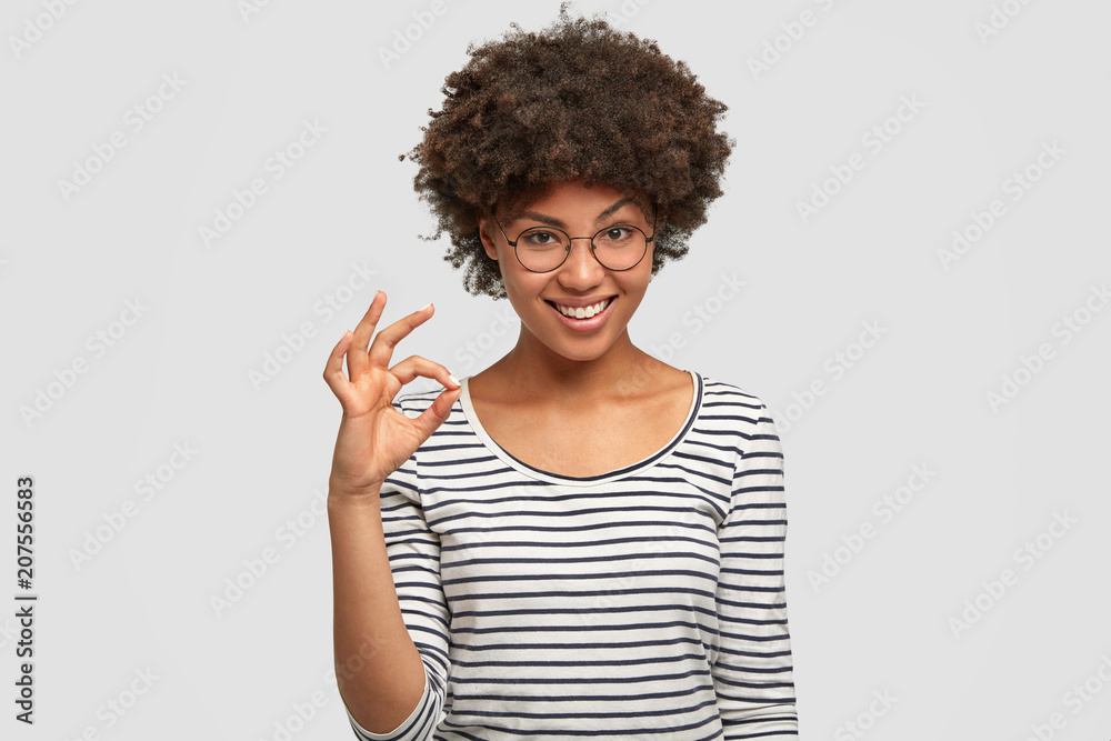 Beautiful mixed race female with Afro hairstyle, demonstrates okay sign, being satisfied with new project, wears casual striped jacket, isolated over white background. Ethnicity, body language concept Beautiful mixed race female with Afro hairstyle, demonstrates okay sign, being satisfied with new project, wears casual striped jacket, isolated over white background. Ethnicity, body language concept