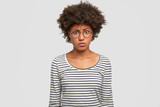 Discontent African American female has crisp dark hair, frowns face and looks puzzled, has angry expression, doesn`t like prepared dish by husband, wears striped sweater. Negative emotions and feeling