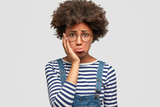 Dull doleful young African American female has sorrowful expression, purses lips and keeps hand on cheek, being abused by her husband, finds out about betrayal, isolated over white background