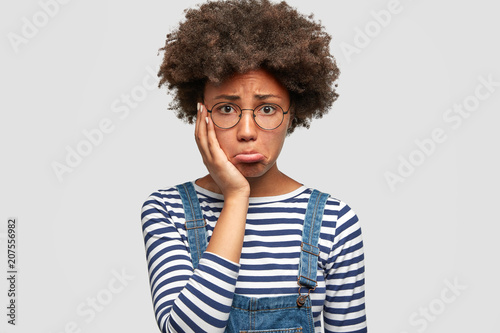 Dull doleful young African American female has sorrowful expression  purses lips and keeps hand on cheek  being abused by her husband  finds out about betrayal  isolated over white background