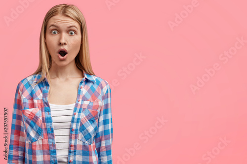 Surprised blonde female student being astonished with bad results of exam, keeps mouth wide opened, wears fashionable checkered shirt, poses against pink blank wall with free space for information