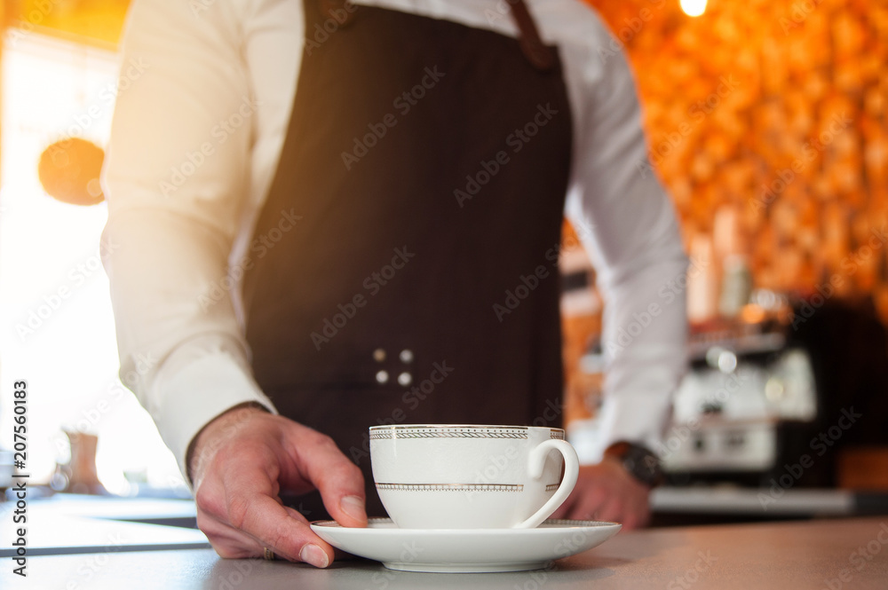 Fototapeta premium Cropped image of handsome barista in apron holding a cup of coffee at the bar counter in cafe. Cup in focus
