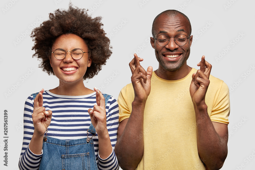 Shot of cheerful hopeful dark skinned female and male students cross fingers up, have desire to pass exam successfully or anticipate results, smile positively. African American guy and his girlfriend Shot of cheerful hopeful dark skinned female and male students cross fingers up, have desire to pass exam successfully or anticipate results, smile positively. African American guy and his girlfriend