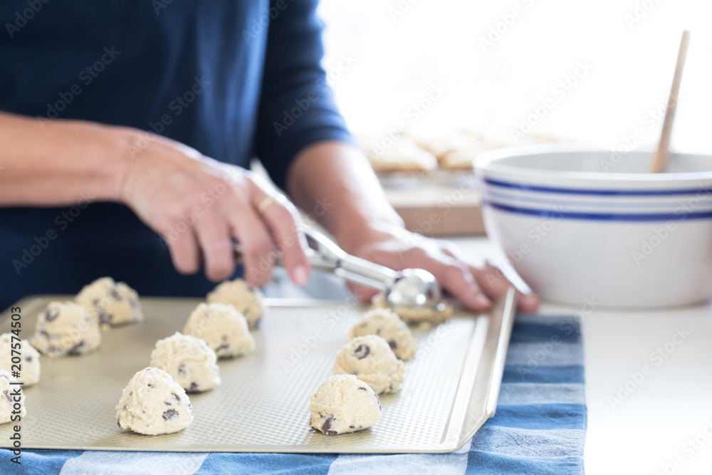 Photograph of a woman’s hands scooping out chocolate chip cookie dough on to a cookie sheet in the kitchen 