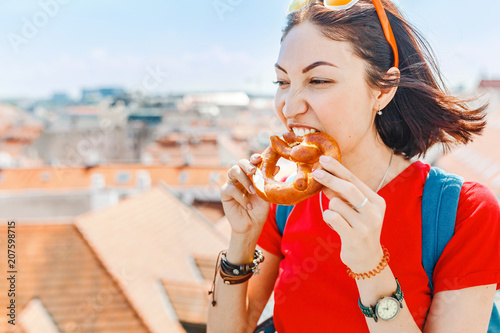 Young female tourist eating traditional bread called pretzel on the historical city center background in Europe