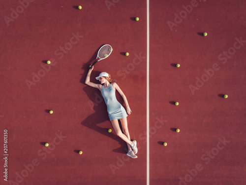 Overhead top view of young Caucasian teen model wearing fashionable tennis dress, lying on tennis hardcourt with a lot of balls, summer sunny day outdoors. Fashion portrait shoot © supamotion