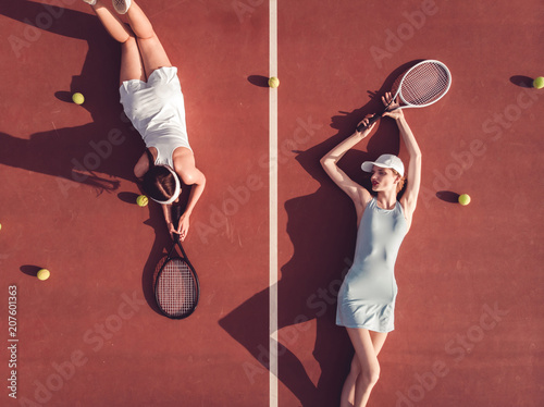 Overhead top view of two young Caucasian teen models wearing fashionable tennis dresses, lying on tennis hardcourt with a lot of balls, summer sunny day outdoors. Fashion portrait shoot © supamotion