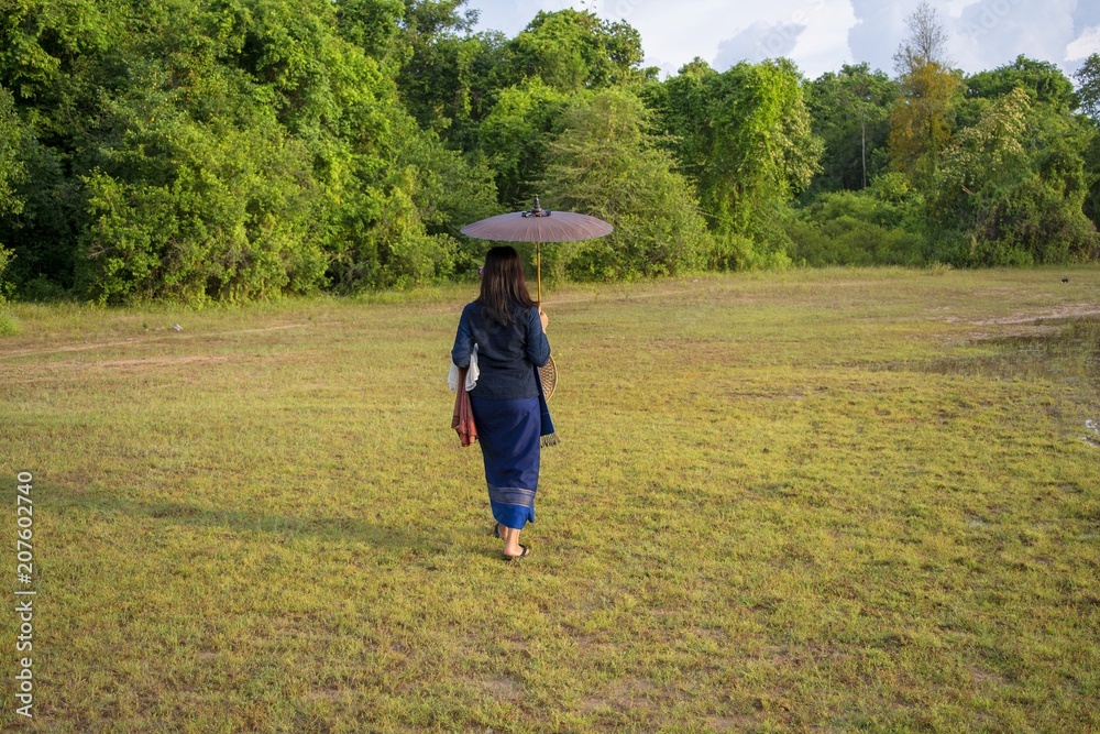 Asian women walking on umbrellas,Thai women walk in the grass./Thai women in blue sarongs are walking past the grass and have a backdrop of forest. Asian women walking on umbrellas,Thai women walk in the grass./Thai women in blue sarongs are walking past the grass and have a backdrop of forest.