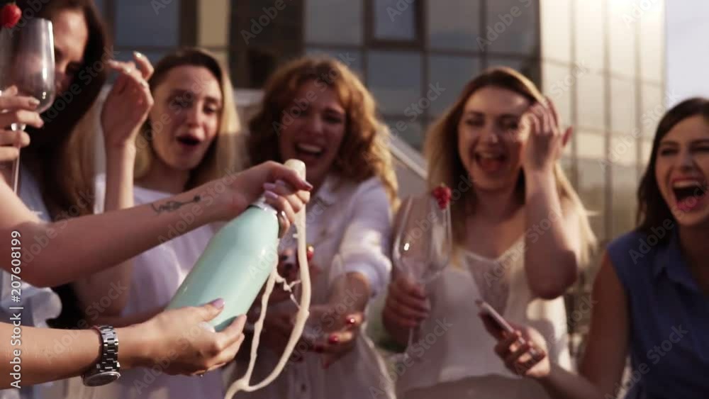Beautiful young ladies celebrate. Hen-party. Shaking a bizarre bottle of champagne and letting it spraying. The girl pours a drink into glasses. Outside. Glamorous party. Slow motion Beautiful young ladies celebrate. Hen-party. Shaking a bizarre bottle of champagne and letting it spraying. The girl pours a drink into glasses. Outside. Glamorous party. Slow motion
