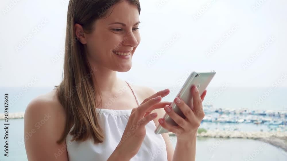 Smiling young woman in white dress using mobile on balcony. Female on vacation enjoying sea view from terrace in morning and talking with friend via cellphone messenger app in slow motion Smiling young woman in white dress using mobile on balcony. Female on vacation enjoying sea view from terrace in morning and talking with friend via cellphone messenger app in slow motion