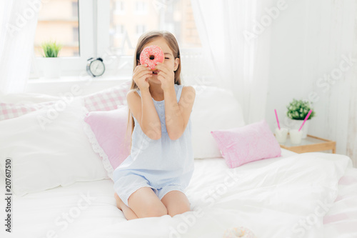 Small pretty female child holds tasty sweet doughnut, poses in bedroom on comfortabled bed, dressed in pyjamas, going to have breakfast, has joy and poses at camera. Chilhood and bedding concept
