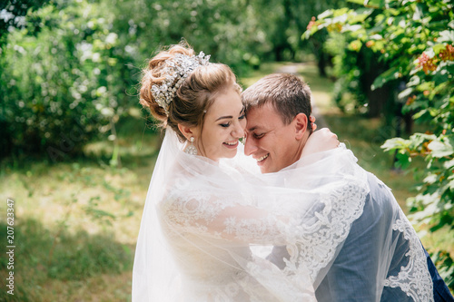Portrait of the bride and groom hugging and laughing in the park. A girl in a white lace veil embraces her man and steals it with her.