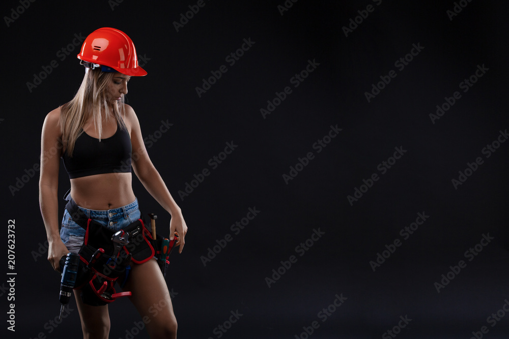 attractive sexy girl in safety helmet with tool belt holding electric drill on black background. Copy space. attractive sexy girl in safety helmet with tool belt holding electric drill on black background. Copy space.