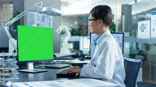 Asian Scientist Sitting at His Desk Works on a Personal Computer with Mock-up Green Screen. In the Background Computer Science Research Laboratory with Robotic Arm Model.