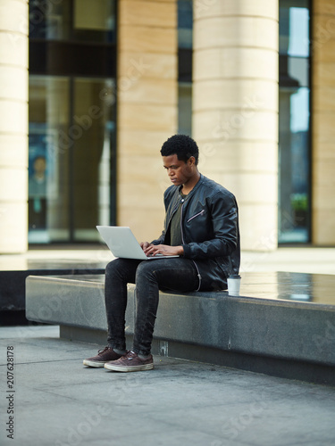 Young African American man sitting on stone street bench and working on laptop outdoors, paper coffee cup near him