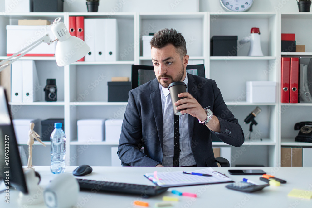 A man sits in the office at the table, drinks coffee and looks at the computer monitor. A man sits in the office at the table, drinks coffee and looks at the computer monitor.