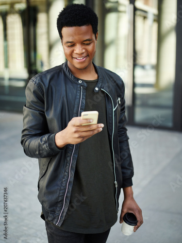 Smiling African boy walking down street with coffee cup in his hand and texting on cell phone