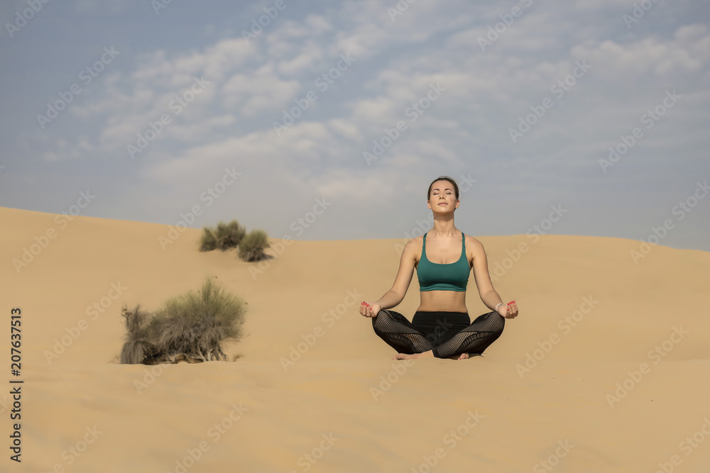 Slim athletic white young woman performs yoga pose in the desert wearing long black tights and a green sports bra 