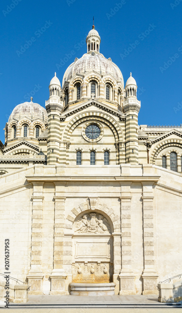 The cut stone fountain of the double flight stairway leading up to the cathedral of Sainte-Marie-Majeure in Marseille, with the cupolas, chapels and turrets of the religious building against blue sky.