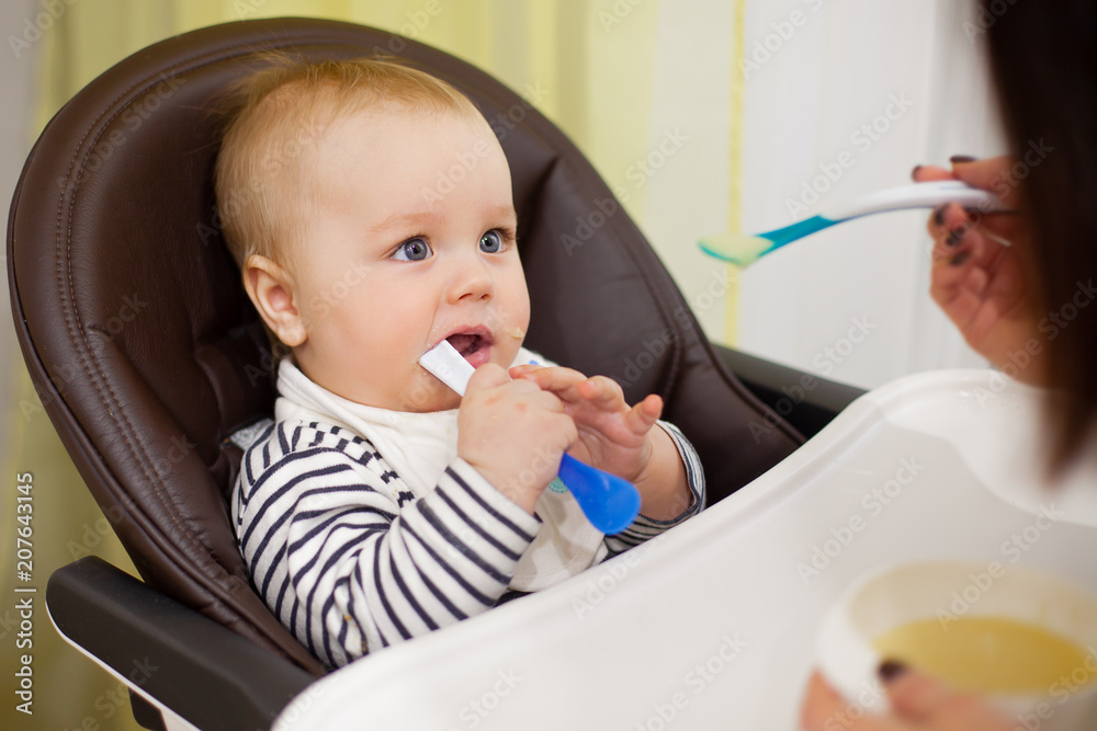 Young mother feeding her little baby son with porridge,who sitting in high baby chair for feeding Young mother feeding her little baby son with porridge,who sitting in high baby chair for feeding
